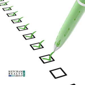 A close-up of a green pen checking off boxes on a list, symbolizing the steps in the home-buying process. The Ketcham Realty Group logo is in the bottom left corner