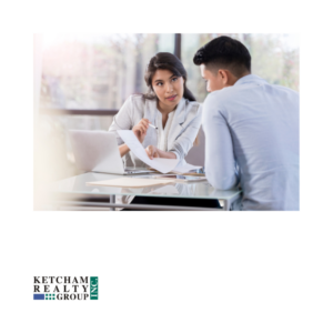 A professional real estate agent sitting across from a client, reviewing important documents related to a home purchase. The Ketcham Realty Group logo appears in the lower-left corner.